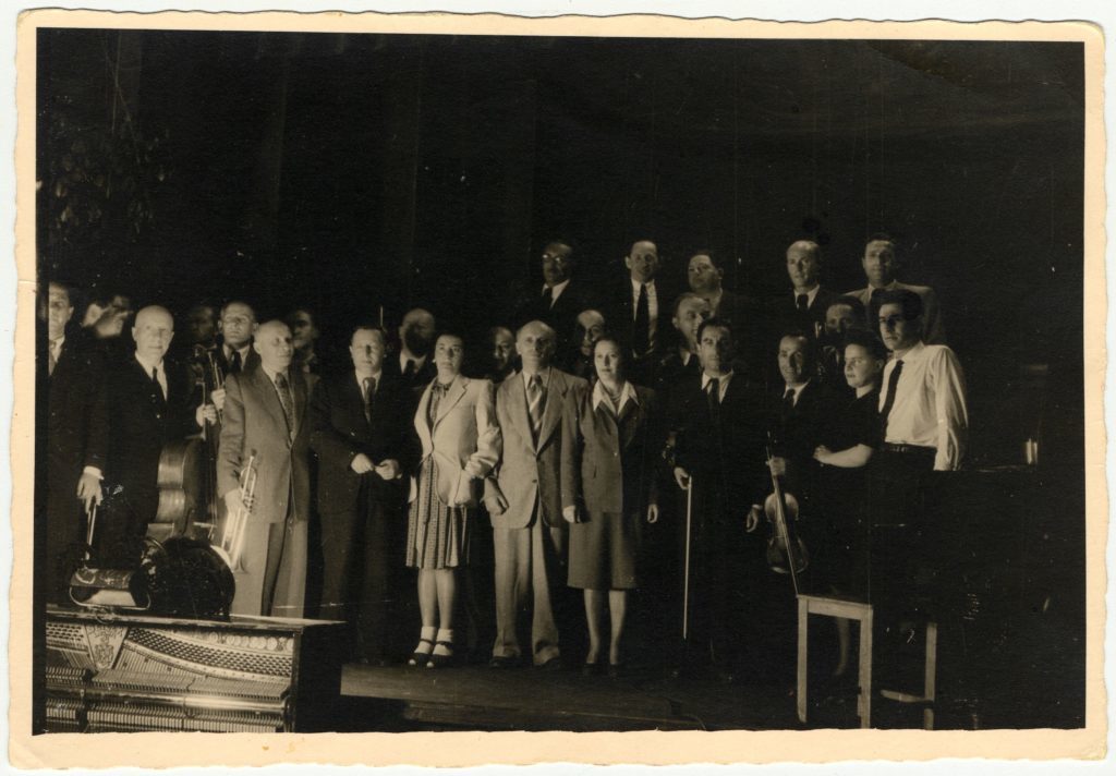 Performance of the Ex-Concentration Camp Orchestra conducted by Leonard Bernstein. Bernstein conducted two concerts the same day--a matinee in Feldafing followed by an evening concert in Landsberg. It is unclear in which camp this photo was taken. Pictured are Leonard Bernstein (far right), Fania Durmashkin (to his left), Max Beker (fourth from the right) and Henia Durmashkin (seventh from the right). (Credit: United States Holocaust Memorial Museum, courtesy of Sonia Beker)
